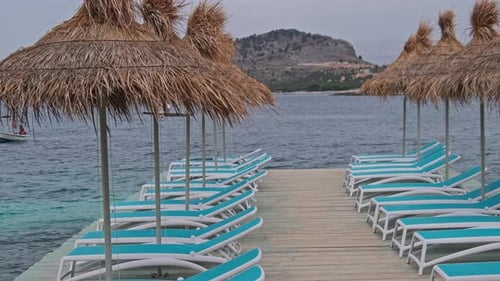 Empty Sun Loungers with Straw Umbrellas in Row on Pier By Beach in Turquoise Sea