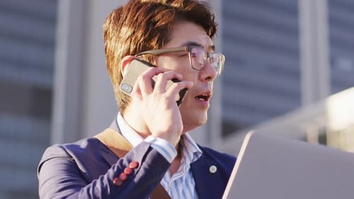 Asian man talking on smartphone while using laptop at corporate park