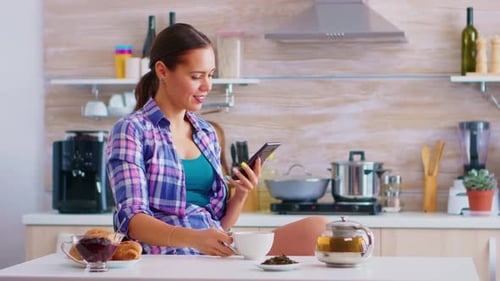 Woman Using Mobile Phone While Drinking Tea in Kitchen