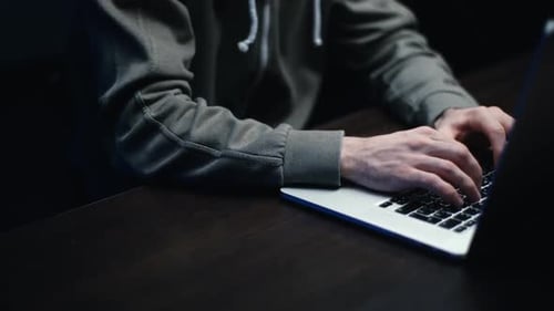 Man in Hoodie Typing on Laptop in the Dark