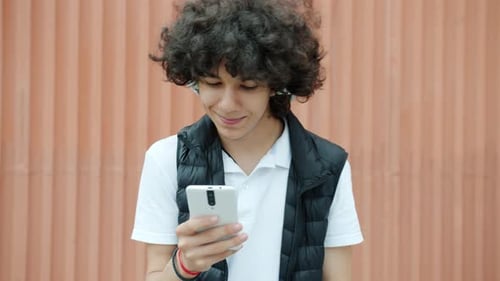 Cheerful Mixed Race Teenage Boy Wearing Headphones Using Smartphone in the Street Having Fun