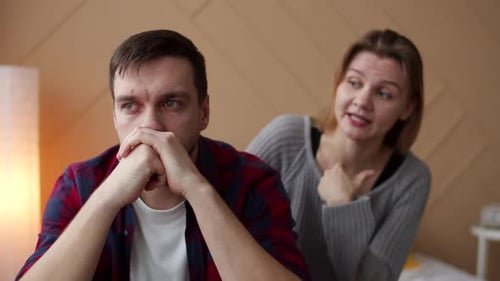 Man Face Red From Tears Sitting on Bed While Wife Swears at Him Front View
