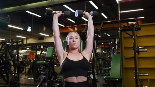 Woman lifts dumbbells training her shoulder muscles in gym. Hard workout