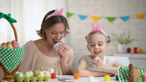 Schöne Mutter und Tochter in lustigen Stirnbändern essen Osterschokoladeneier