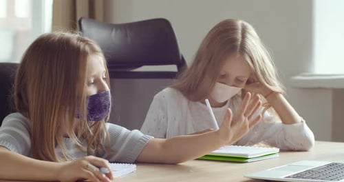 Two Schoolgirls in Medical Protective Masks Sitting Together at Table at Home at School Classroom