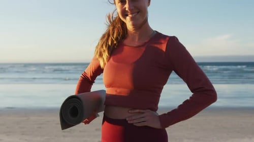 Portrait of caucasian woman holding yoga mat at the beach smiling