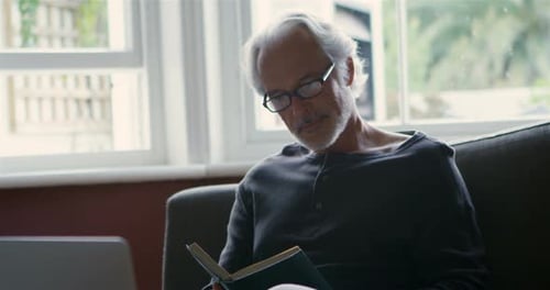 Senior Man Reads Book on Sofa Indoors
