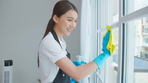 Asiatische Putzfrau putzt im Wohnzimmer, wischte unordentliches schmutziges Glasfenster zu Hause ab.