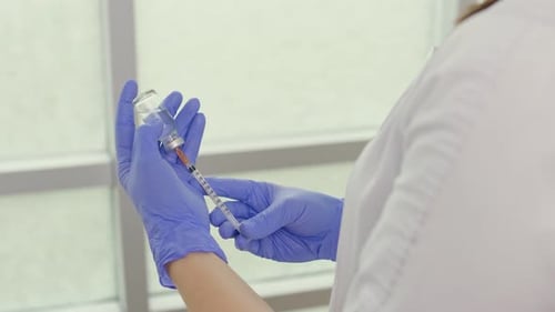 A Doctor in a Medical Mask and Blue Gloves Holds a Syringe and a Vaccine