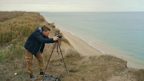 Man Adjusting Camera on Tripod Near Ocean Coastline