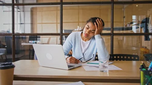 Müde Afroamerikanerin sitzt am Schreibtisch im Büro und hält Hand auf Kopf und nimmt sie ab