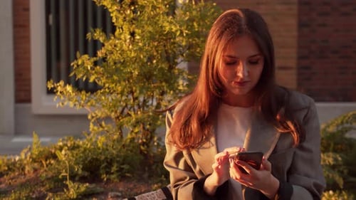 Young attractive business woman sitting outdoor on the bench and using smartphone.