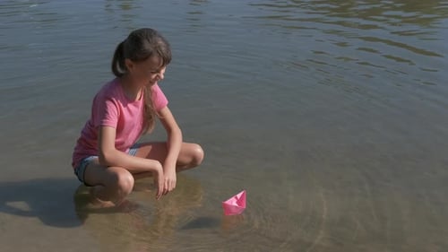 Girl Plays with Pink Paper Boat on Shore