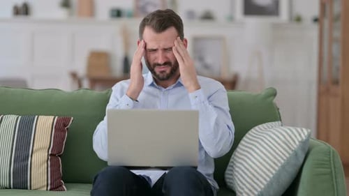 Stressed Man with Headache Using Laptop on Couch