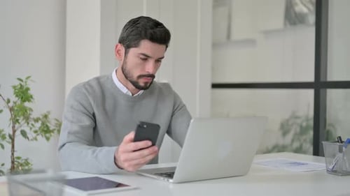 Young Man Using Smartphone While Using Laptop in Office