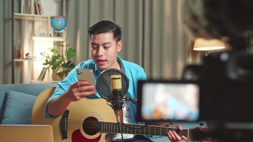 Un hombre asiático es un vlogger con guitarra Lee un comentario en un teléfono móvil mientras transmite en vivo