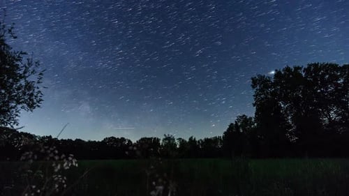 moving milky way and star trails on night sky over rural landscape with silhouette of trees