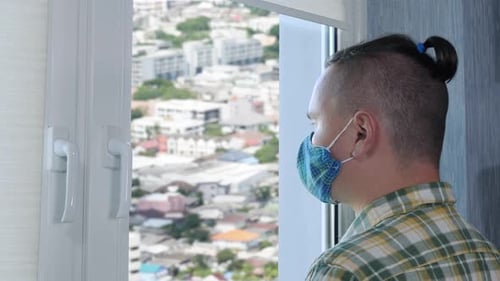 Homem adulto triste com máscara protetora olha pela janela durante a quarentena