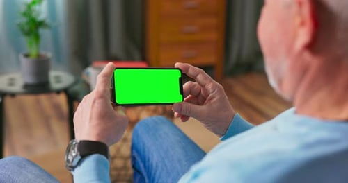 Homem mais velho em casa descansando em um sofá usando com telefone inteligente de tela de maquete verde