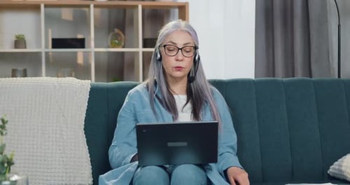 Mujer mayor con auriculares sentada frente a la computadora portátil durante la videoconferencia