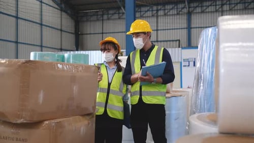 Two Asian factory or warehouse workers stand among stack of products in workplace