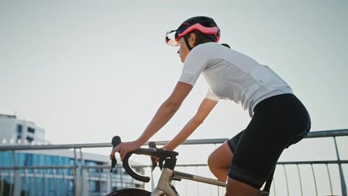 Woman Professional Cyclist in Protective Helmet is Riding Her Bike Uphill Along Bridge Surrounded By