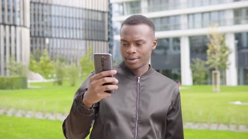 A Young Black Man Has a Video Call on a Smartphone, Waves His Hand and Smiles - Office Buildings