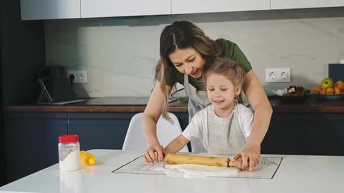 Mother and Little Daughter Roll Dough at Table in Kitchen