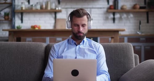 Focused Young Man in Headphones Works on a Laptop Holding It on His Lap and Sitting on a Sofa at
