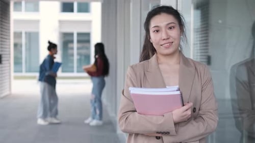 Female university student holding folders and looking at the camera while standing at the hallway