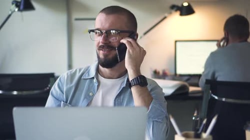 Young Businessman is Talking on Phone at Table with Laptop in White Office