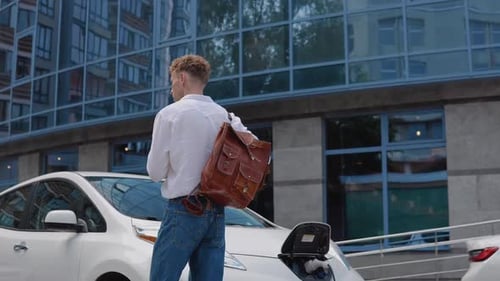 Stylish Modern Young Curly Man with a Stylish City Briefcase on His Shoulder Approaches His Electric