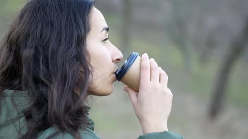 On a cloudy day, a beautiful young woman in a park holds a glass in his hands and drinks coffee