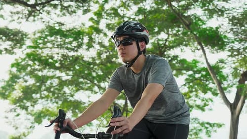 Asiatischer junger aktiver Mann in Sportkleidung, der abends im öffentlichen Park aus Gesundheitsgründen Fahrrad fährt.
