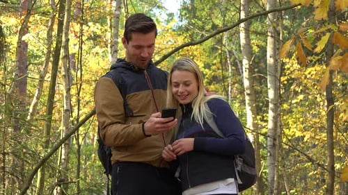 A Hiking Couple Stands in a Forest on a Sunny Day, Woman Takes a Picture of Something Off Camera