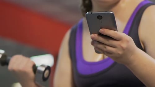 Woman Using Phone While Lifting Dumbbell at Gym