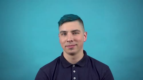 A Young Man in Glasses with Blue Hair Winks at His Eyes. Emotional Man Winks on a Blue Background.