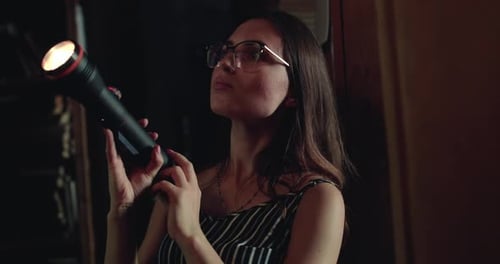 Girl in Glasses with a Lantern Views the Library Shelves in the Darkness