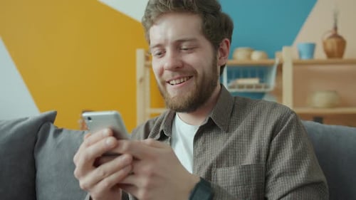 Man Smiling Using a Smartphone on Couch Indoors