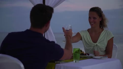 A man and woman clink their wine glasses with dinner on a tropical island beach