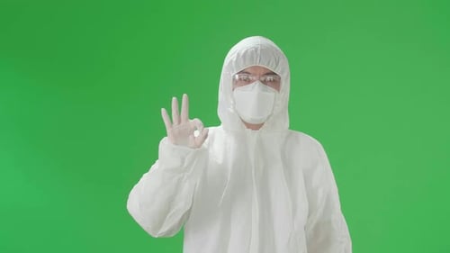 Man Wear Protective Uniform PPE, Medical Face Mask And Showing Hand Ok Sign In Green Screen Studio