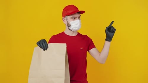 Junger Zusteller mit medizinischer Schutzmaske und Handschuhen hält Paket und zielt zur Seite