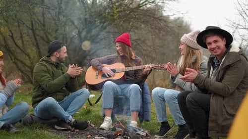 Group of Friends camping.They Are Sitting Around Camp Fire, Playing Guitar
