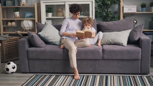 Young Woman Reading Book To Her Son Sitting on Sofa in Studio Apartment Together