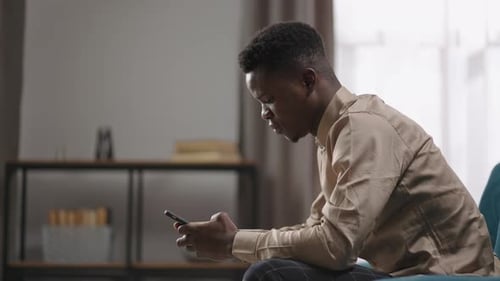 Afroamerican Student is Using Smartphone Sitting Alone at Home Communicating in Social Nets Male