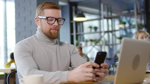 Cheerful Caucasian Man Texting on Phone and Smiling for Camera in Cafe