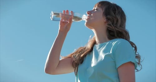 Woman of European Appearance in the Open Air Drinks Water From a Bottle Against a Clear Blue Sky