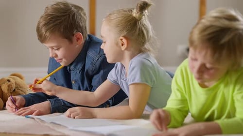 Side View of Joyful Caucasian Children Drawing Lying in Living Room at Home