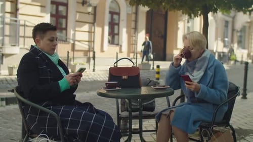 Mature Businesswomen Sitting at Cafe Table Using Smartphone Outdoors
