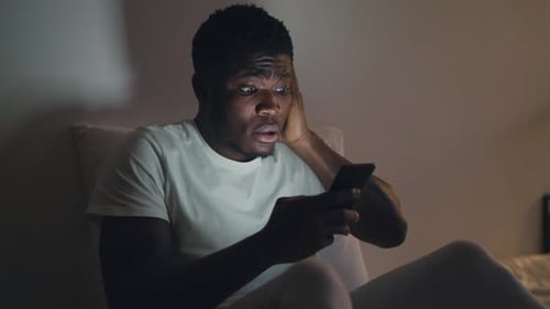 Junger besorgter Afroamerikaner, der Nachrichten auf dem Smartphone liest, spät zu Hause auf dem Smartphone surft